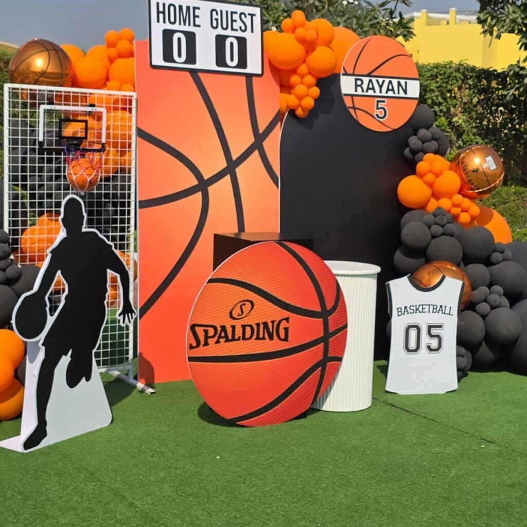 Basketball party decorations with orange balloons, a scoreboard, and basketball cutouts on grass.