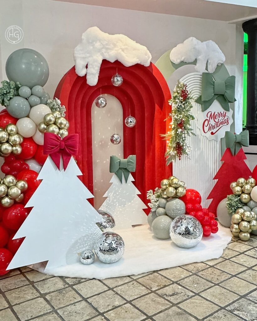 Christmas party backdrop with red arches, white tree cutouts, and gold balloons.