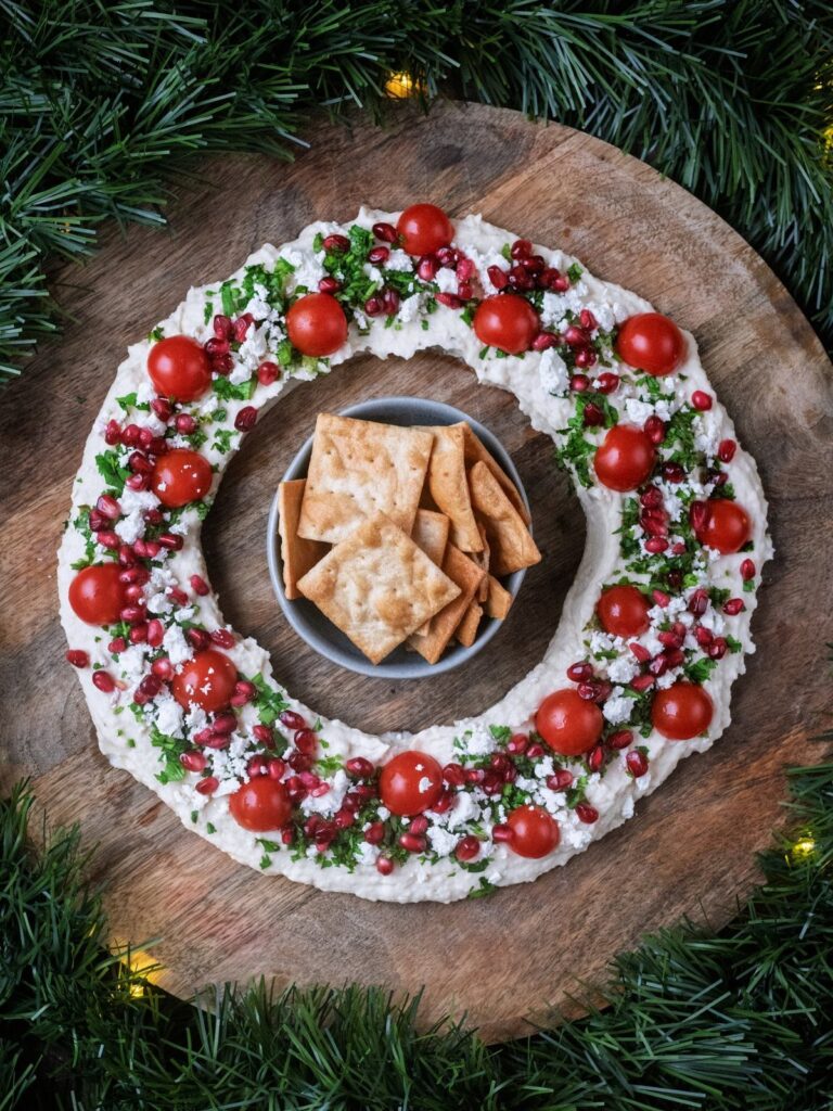 Wreath-shaped creamy dip with tomatoes and pomegranate seeds served with crackers on wood.