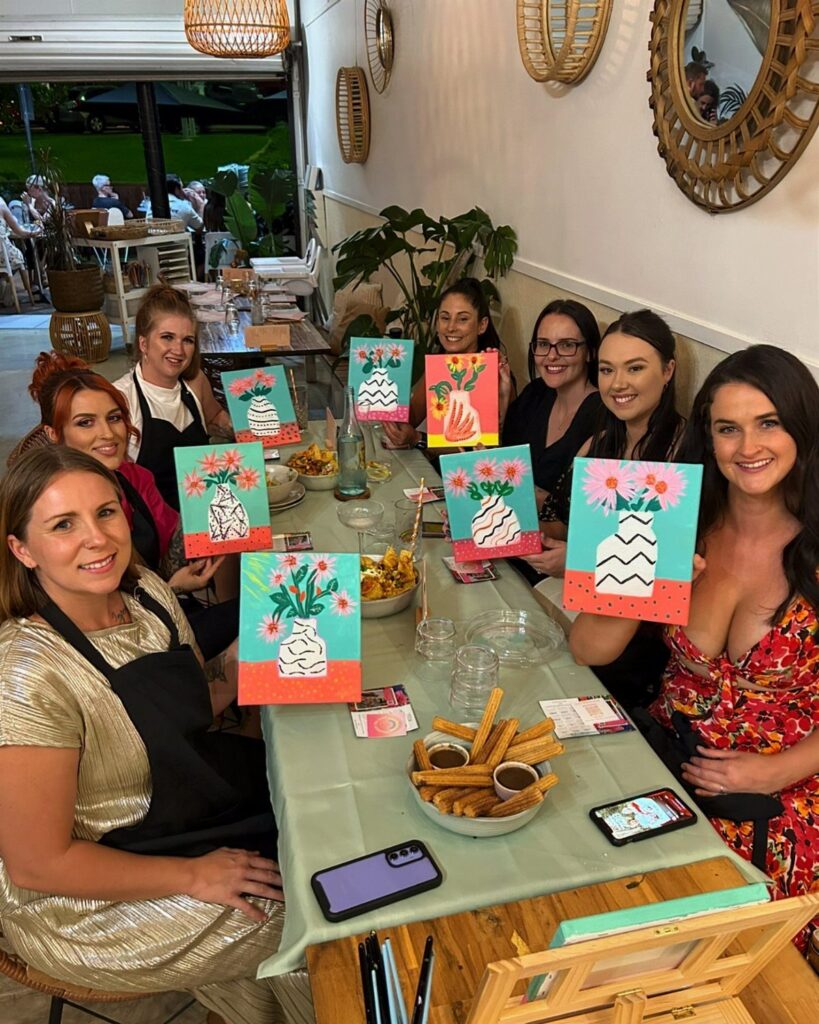 Women holding painted flower canvases while sitting around a table with snacks.