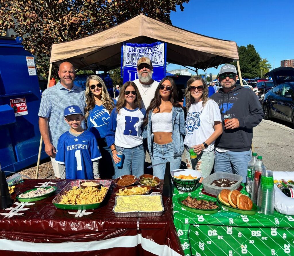 Group of people posing behind a food spread at a football tailgate party.