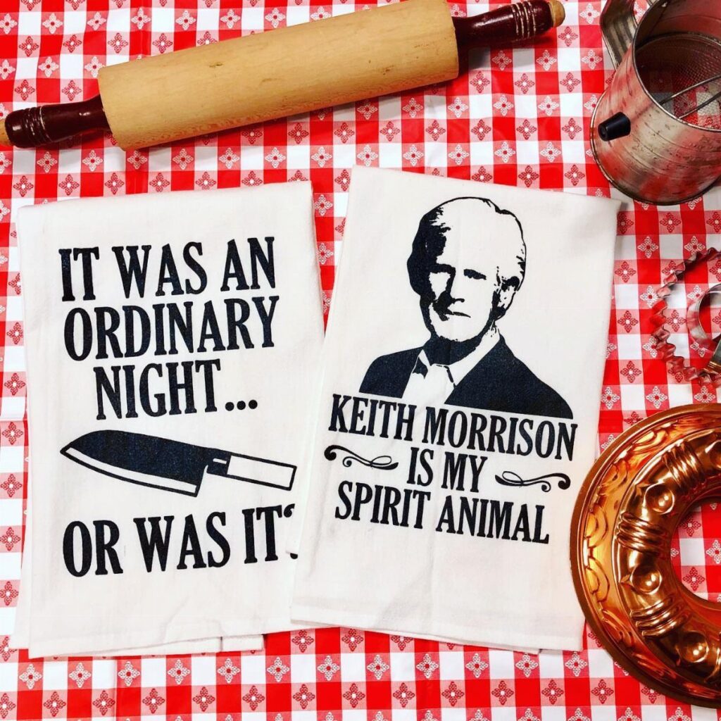 Two white humorous kitchen towels on a red checkered tablecloth with a rolling pin.