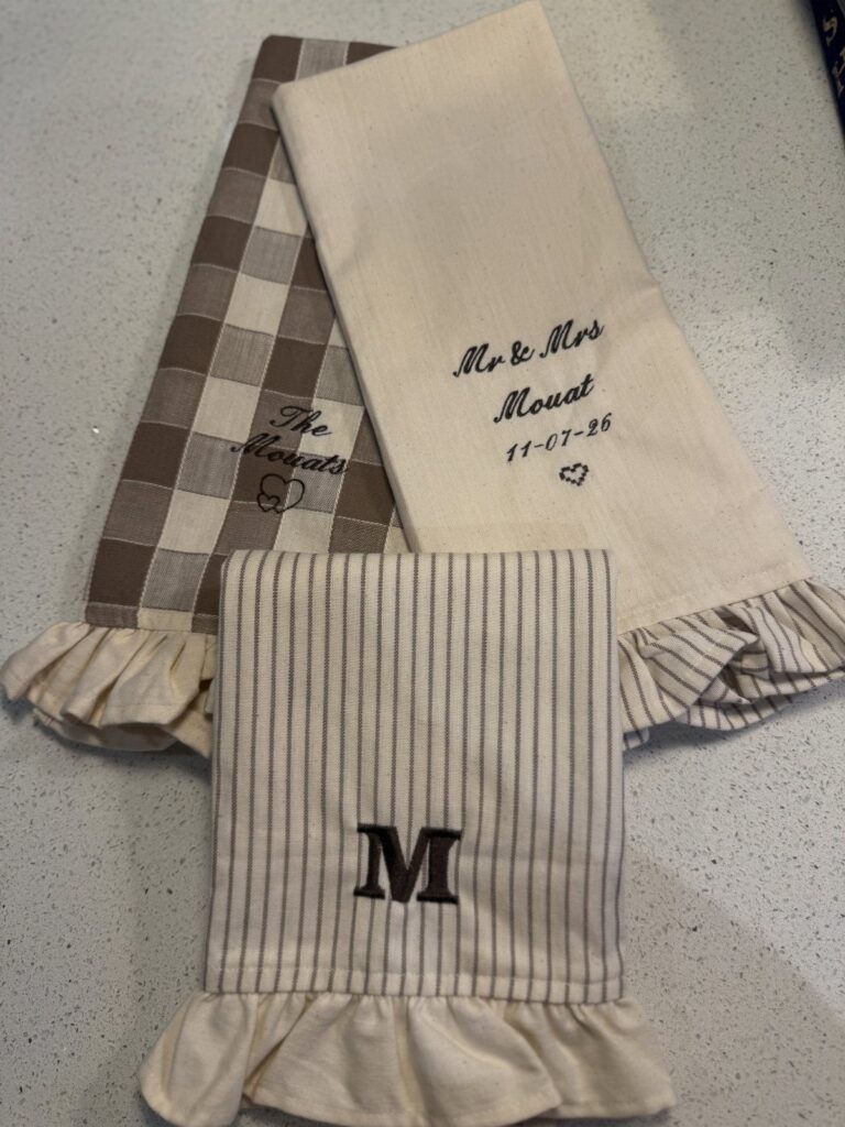 Three personalized kitchen towels with ruffles and embroidery in neutral tones on white.
