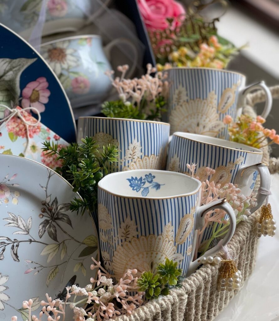 Woven gift basket with blue striped mugs, floral plates, and decorative artificial flowers.