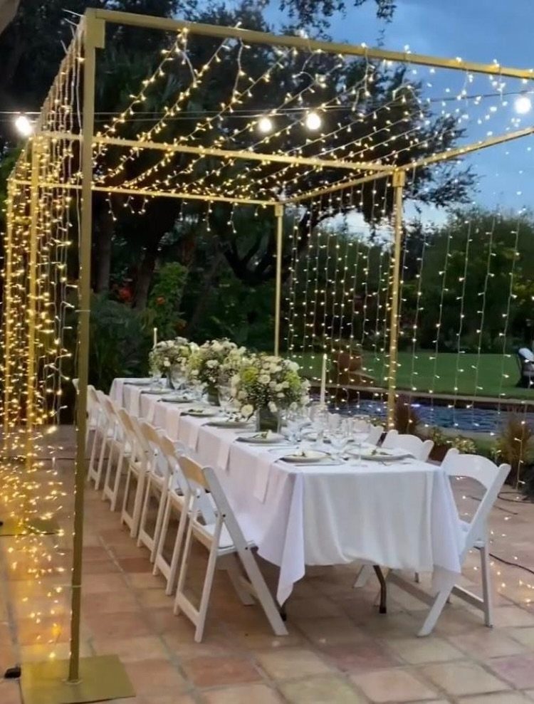 Outdoor dining table with white linens and flowers under a gold fairy light canopy