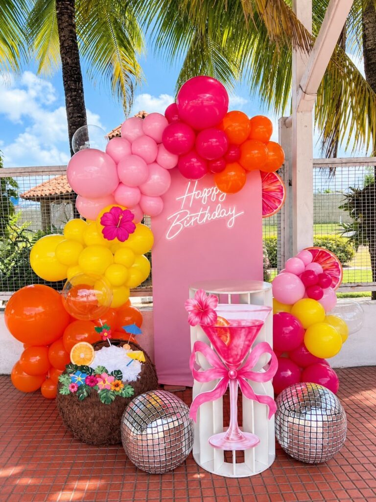 Colorful balloon arches and tropical decorations surrounding a pink Happy Birthday backdrop outdoors.