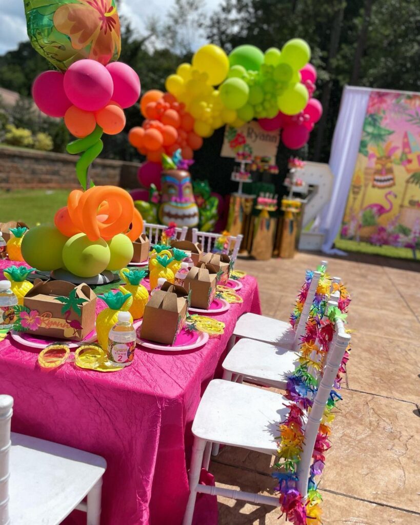 Tropical party table with pink tablecloth, colorful balloons, and vibrant ribbon-decorated chairs.