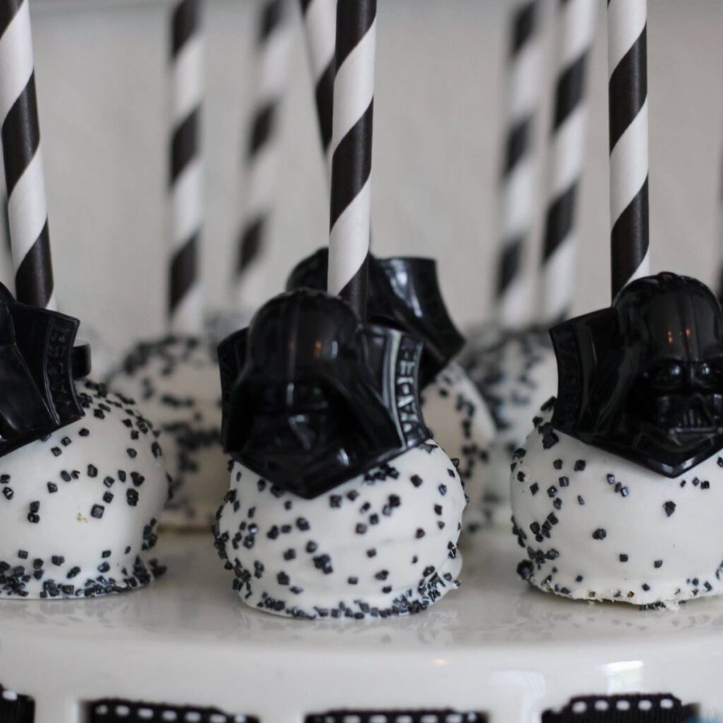 White cake pops with black sprinkles and Darth Vader helmet toppers on striped straws.