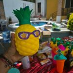 Pineapple shaped drink container with sunglasses held in front of a pool party