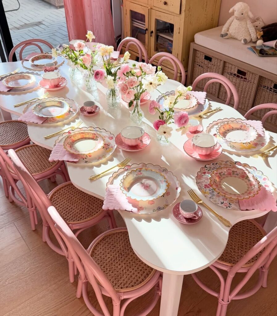 Tea party table setting with pink chairs, floral plates, and fresh pink flowers.