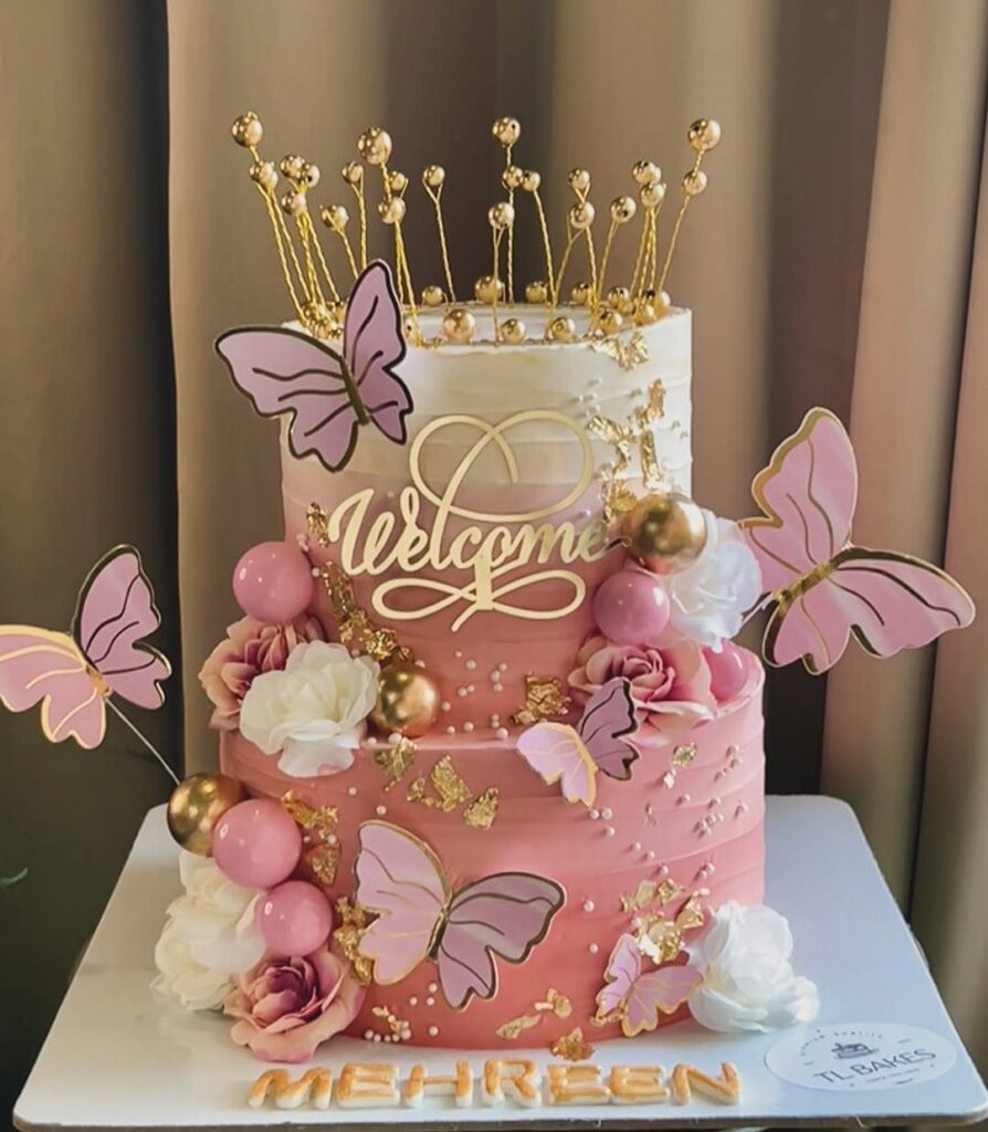 Three-tier pink and white cake with gold crown, butterflies, and Welcome topper.