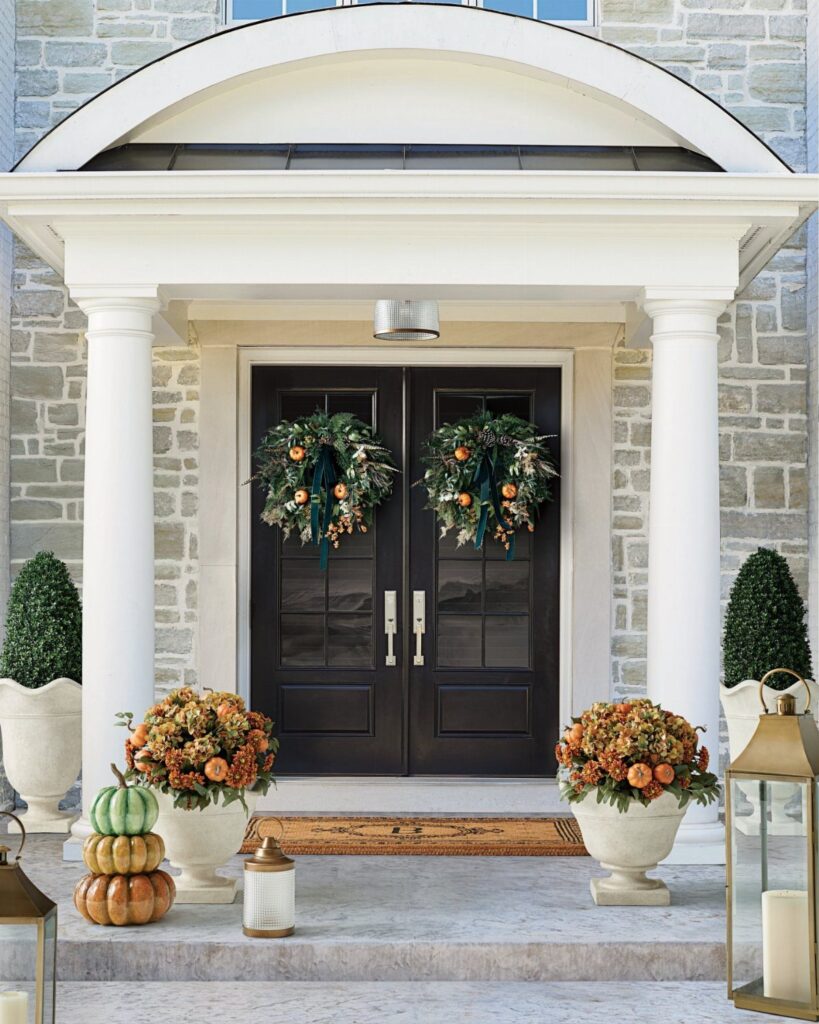 Two autumn wreaths on black double doors with pumpkins and flowers on the porch.